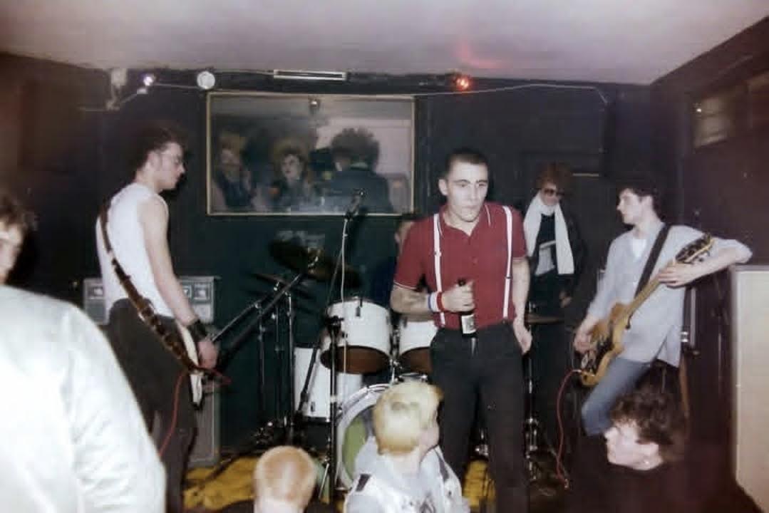 Band playing a small dark club room, singer in red shirt with braces, crowd right at the stage